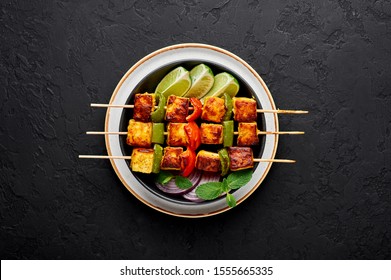 Paneer Tikka at skewers in black bowl at dark slate background. Paneer tikka is an indian cuisine dish with grilled paneer cheese with vegetables and spices. Indian food. Top view