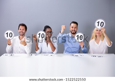 Panel Of Excited Judges Sitting In Front Of Grey Wall Showing 10 Score Cards