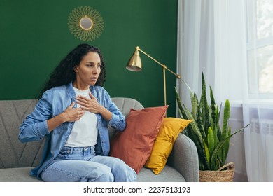 Panic attack woman alone at home scared hispanic woman having trouble breathing sitting on couch in living room.