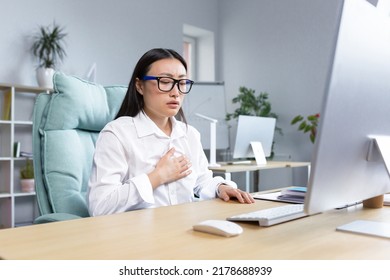 Panic Attack In The Workplace. Young Asian Woman Sitting At Desk In Office, Holding Chest, Breathing Hard, Panic Attack, Trying To Calm Down.