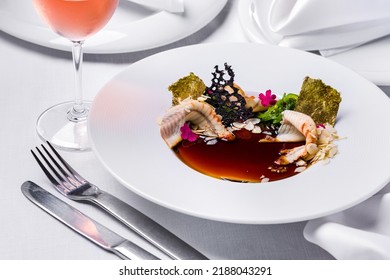 Panna Cotta With Smoked Eel And Balsamic Sauce, Top With Chuka Salad, Almond Flakes, Edible Petals, Gilded Nori Seaweed. The Food Is In A Deep White Plate. The Plate Is On A White Tablecloth, Cutlery 