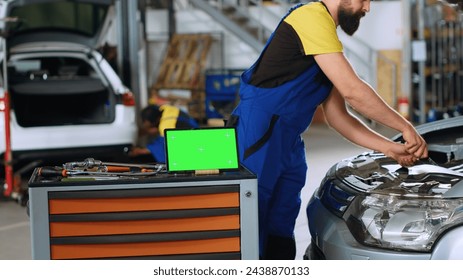 Panning shot of green screen laptop in garage workspace, sitting on work bench next to professional tools with mechanics in background working on cars, using torque wrench to fix them - Powered by Shutterstock