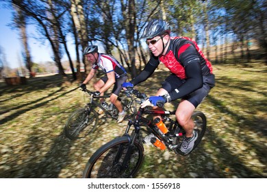 Panning Shot Of Two Mountain Bikers, Racing In A Forest.  Movement, Some Of The Bikers In Focus.