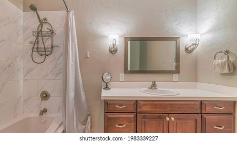 Pano Sink over cabinets and built in bathtub with shower inside bathroom of a home. Small mirror, wall lights, white curtain, and tiles can also be seen inside the wash room.
