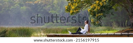 Panorama of asian woman sitting on the edge of dock with peaceful natural park during summer with yellow flower blossom for serene and relaxation outdoor recreation