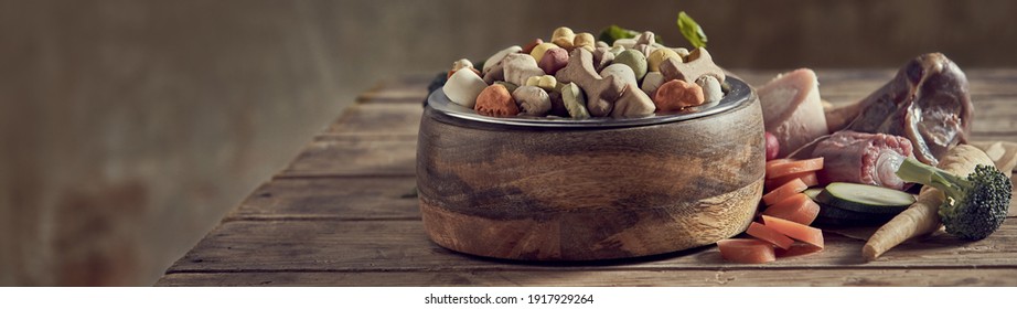 Panorama-Banner für gesunde, ausgewogene Lebensmittel für Ihren Haushund mit einer Schüssel gefüllt mit getrockneten Keksen, umgeben von frischen pflanzlichen Zutaten und Knochen mit Kopienraum für Werbung