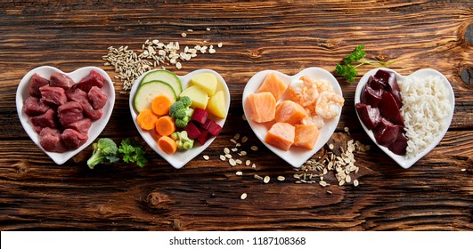 Panorama banner of healthy fresh ingredients for pet food in individual heart-shaped bowls viewed from overhead with chopped raw beef, liver and chicken , mixed vegetables and rains on rustic wood - Shutterstock ID 1187108368