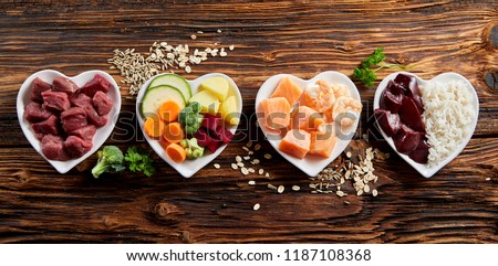Panorama banner of healthy fresh ingredients for pet food in individual heart-shaped bowls viewed from overhead with chopped raw beef, liver and chicken , mixed vegetables and rains on rustic wood
