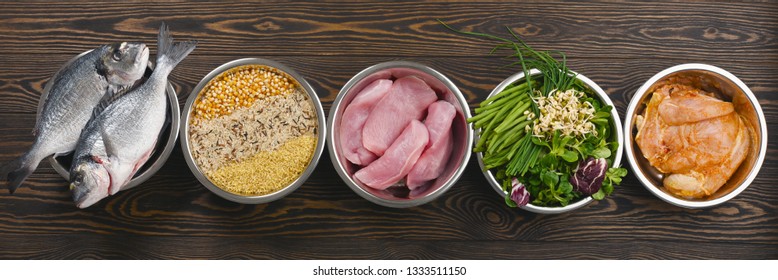 Panorama banner with healthy pet food ingredients with chopped raw turkey, fish, groats, greens and grains in individual bowls on brown wooden background. Flat lay.