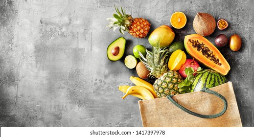 Panorama Banner Of A Large Selection Of Healthy Organic Tropical Summer Fruit Spilling From A Reusable Grocery Bag Onto Texture Grey Slate With Copy Space Viewed From Overhead