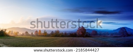 panorama of a carpathian rural landscape at twilight. arable in front of a forest in colorful foliage. day and night time change concept. distant mountains beneath a sun and moon. hazy atmosphere