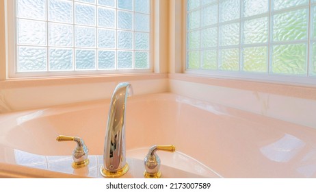 Panorama Corner Bathtub With Deck Mounted Widespread Stainless With Gold Faucet Fixture. Bathtub In A Bathroom Against The Large Windows With Textured Glass Panels.