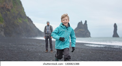 Panorama des süßen positiven Jungen, der am schwarzen Sandstrand von Reynisfjara in Eisland mit schöner Aussicht auf die Felsen von Rynisdrangar und seinen Vater im Hintergrund, Familienaktives Urlaubskonzept