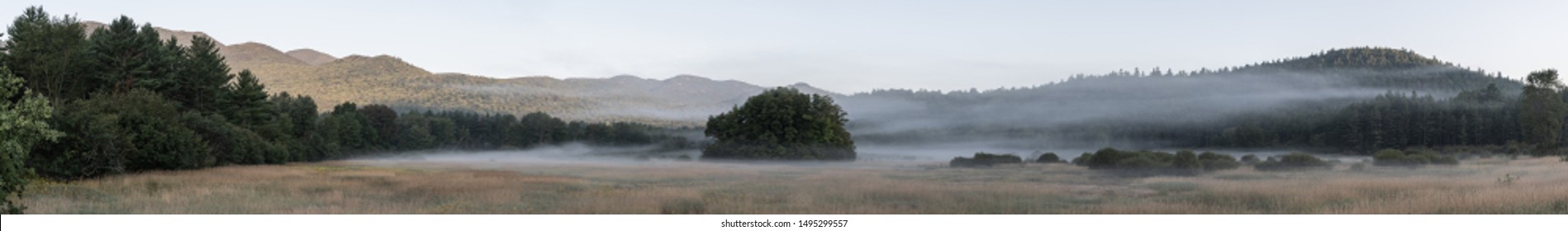 Panorama of early morning mist in the Adirondacks National Park. Field with mountains in the background. Cold August sunrise. Shot in the countryside of Keene (Lake Placid), NY, USA.