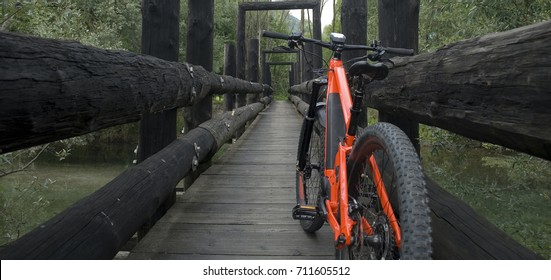 Panorama: Ebike E-bike electric bicycle orange, leaning on an old dark wooden bridge, nestled in the woods, under which flows a small light green mountain river, Ossola, Alps, Italy