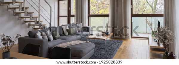 Panorama of elegant, gray corner sofa with\
decorative pillows in spacious living room with big windows and\
stairs to second floor
