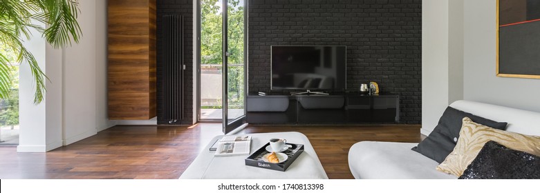 Panorama Of Elegant Living Room With Tv On Black Brick Wall, Wooden Floor And Balcony Door