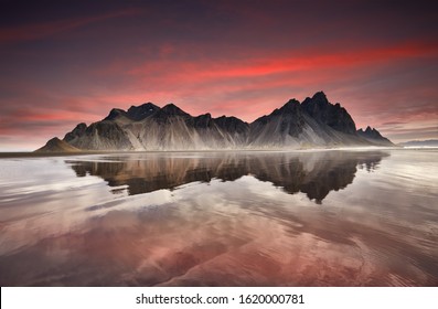 Panorama of Famous Stokksnes mountain reflected in water on Vestrahorn cape (Batman Mountain), Iceland, Europe. Popular tourist attraction. Beauty world. Red sky over black sand dunes on the beach. 
