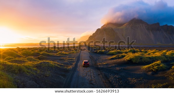Panorama\
of a gravel road at a golden Sunset with Vestrahorn mountain in the\
background and a car driving the road in\
Iceland