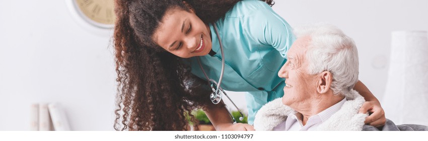 Panorama of a happy nurse with a stethoscope covering an elderly man with a blanket in a nursing home