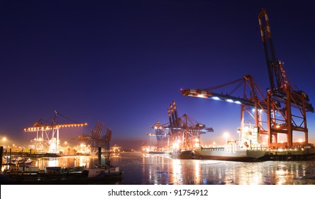 Panorama image of the illuminated cargo port in Hamburg at night with container terminals, cargo ships and cranes and a clear blue sky.