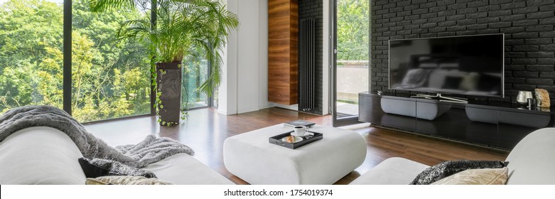 Panorama Of Living Room With Tv On Black Brick Wall, Ottoman For Coffee Table And Big Windows