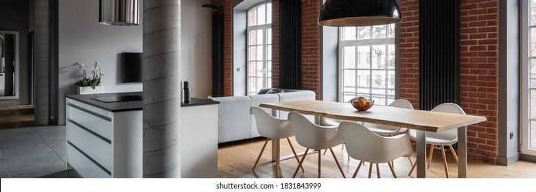 Panorama Of Modern Kitchen In Loft Apartment With Industrial Brick Wall And Wooden Dining Table Under Black Pendant Lamp