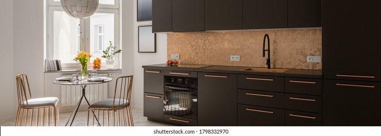 Panorama of modern kitchen with stylish black cupboards and drawers, copper hexagonal tiles and handles and with new black dining table and two, elegant copper chairs