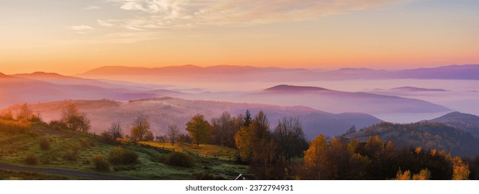 panorama of mountainous countryside landscape at dawn. grassy rural fields on the rolling hills in morning light. trees in fall colors. fog in distant valley. bright sky with clouds above the ridge - Powered by Shutterstock