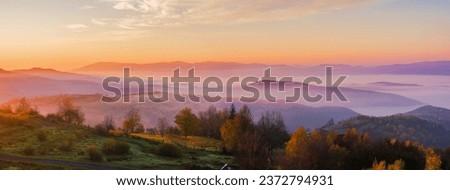 panorama of mountainous countryside landscape at dawn. grassy rural fields on the rolling hills in morning light. trees in fall colors. fog in distant valley. bright sky with clouds above the ridge