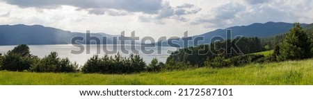Panorama on blue reflecting lake and clear mountain cool water with stone narrow wild shore in evergreen coniferous spruce forest with tall fluffy thorny fir trees, against blue daytime cloudy sky