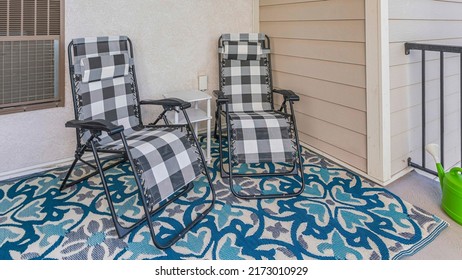 Panorama Porch Of A House With Two Lounge Chairs And Carpet With Blue And White Colors. There Is A Plant Beside The Watering Can On The Left Near The Railings.