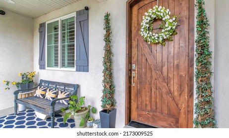 Panorama Porch of a house with wooden front door with digital key access. There are plants at the entrance and bench on a blue carpet near the window with fake shutter design.