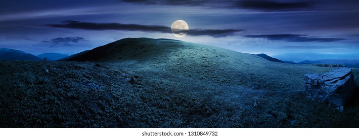 Panorama With Rock On The Grassy Hill In Mountains. Beautiful Summer Landscape In Full Moon Light. Amazing Nature Scenery. Dramatic Cloudy Sky At Night