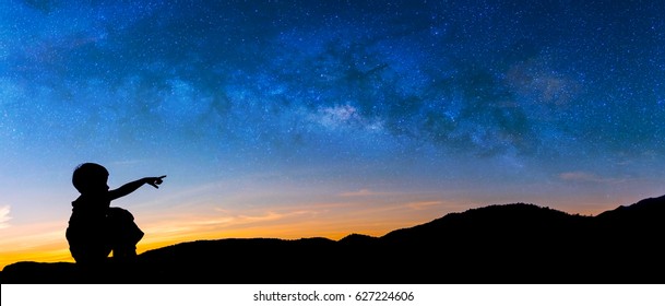 Panorama Silhouette Image Of Happy Boy Point Finger To The Milky Way In Sky With Mountain In Background On Sunset Time.