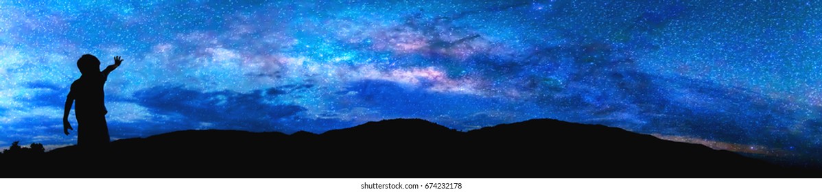 Panorama silhouette image of Happy boy stand and point finger to the milky way on sky with mountain in background on sunset time.