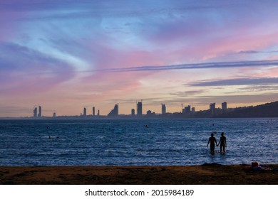 Panorama of the skyline of Limassol. Towering skyscrapers on the horizon, the beach and a couple entering the water in the foreground. Sunset in the sky.