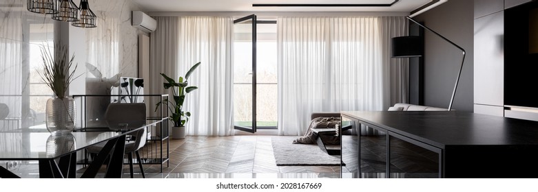 Panorama of spacious and open plan apartment with modern dining table,black kitchen and elegant living room with window wall behind curtains and balcony doors open - Powered by Shutterstock