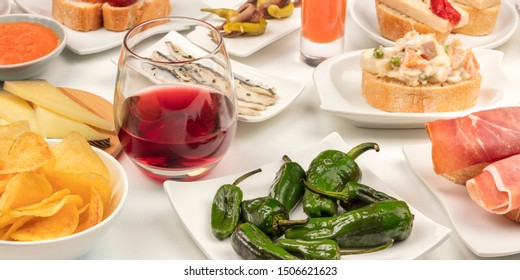 A panorama of Spanish tapas. A glass of red wine with pimientos del padron peppers, ensaladilla rusa salad, jamon, sardines etc, on a white background - Shutterstock ID 1506621623