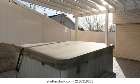 Panorama Sun Flare Covered Portable Spa Pool At The Backyard Of A House. Fenced Backyard With Pergola Roof With String Lights And Stone Tile Flooring Outside The House With Vinyl Wood Siding