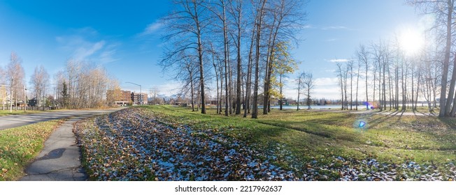 Panorama View Hillside Lakeside Trail With Fallen Brown Leaves Covered In Snow, Dormant Trees Under Sunny Cloud Blue Sky In Lake Spenard, Anchorage. Apartment Complex And Chugach Mountain