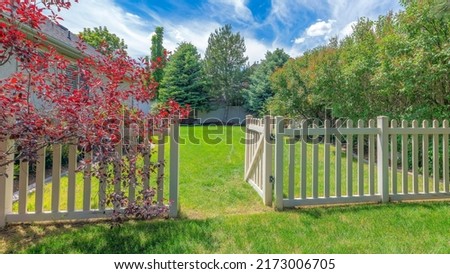 Panorama Whispy white clouds Backyard with picket fence and gate on a green lawn. There are trees on the left near the vinyl fence and a red tree against the house on the right.
