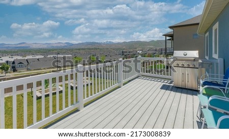 Panorama White puffy clouds Outdoor deck of a house with steel gas barbecue griller and chairs. Wooden deck of a house with white railings and a view of the backyard, residential houses and mountains