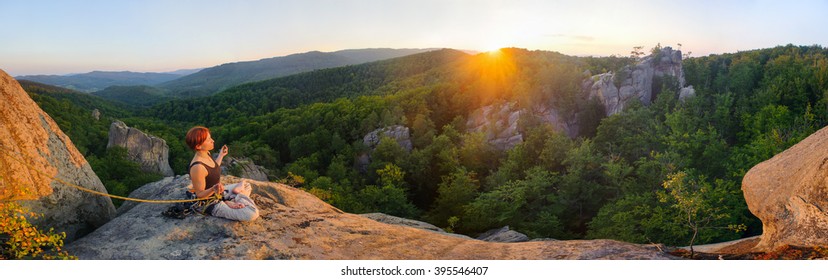 Panorama of young athletic female climber sitting secured with rope and meditating with crossed legs on mountain summit. Beautiful sunset in the mountains.