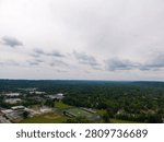 Panoramic aerial view capturing a sprawling suburban town featuring a school campus with a football field and baseball diamond, surrounded by numerous green trees under a cloudy sky