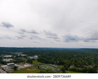 Panoramic aerial view capturing a sprawling suburban town featuring a school campus with a football field and baseball diamond, surrounded by numerous green trees under a cloudy sky
