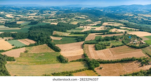 Panoramic aerial view of a patchwork countryside with hay bales, scattered farmhouses, and wooded hills under a summer sky.