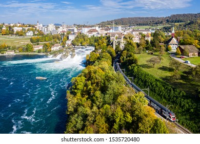 Panoramic aerial view of Rhine Falls and cityscape of Neuhausen am Rheinfall town, Switzerland. Red Swiss train, and tourist boats in waterfall. Cliff-top Schloss Laufen castle, Laufen-Uhwiese.
