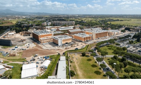 Panoramic aerial view of the Cinecittà Studios complex in Rome, Italy, showing large modern soundstages and production facilities set against the Roman countryside under a bright blue sky.