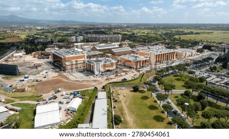 Panoramic aerial view of the Cinecittà Studios complex in Rome, Italy, showing large modern soundstages and production facilities set against the Roman countryside under a bright blue sky.